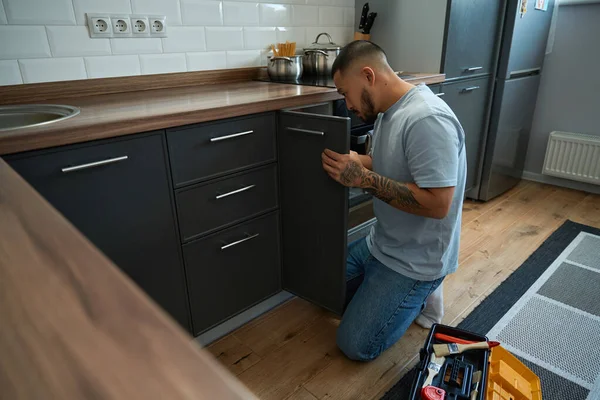 Focused asian man in jeans crouched at door of kitchen cabinet for minor repairs, next to it on floor toolbox