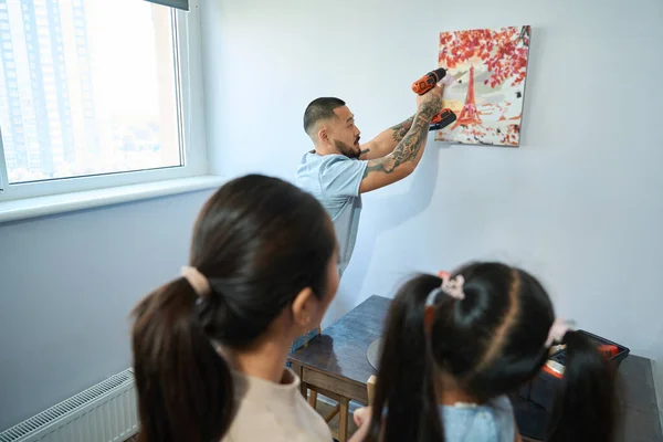 Young man hangs picture over kitchen table with drill in his hands, his wife and daughter look at his work
