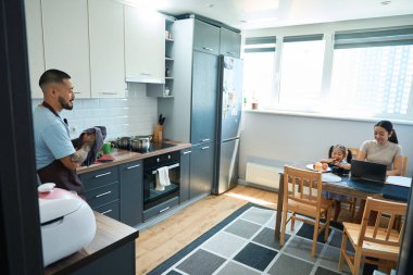 Elinde mutfak havlusuyla gülümseyen bir adam mutfak dolabında duruyor, karısı ve kızı masada oturuyor.