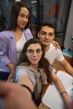 Kendine güvenen bir kadın meslektaşının omzuna eliyle dokunurken grubun üçüncü bir üyesi birlikte selfie çekiyor.