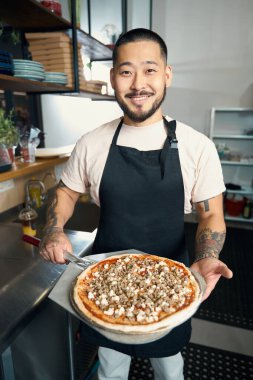 Elinde pizza tutarken kameraya gülümseyen esmer bir genç adam.