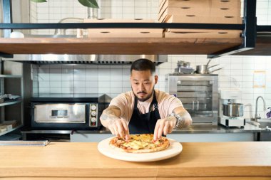 Siyah önlüklü ciddi bir adam yeni pişmiş bir pizzayı süslemeye çalışıyor.