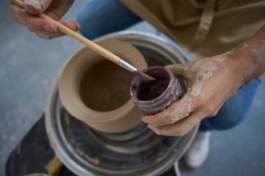 Potter çömlekçi çarkında elinde boya ve elinde fırçayla vazonun üzerinde oturuyor.