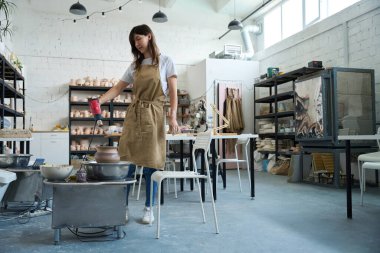 Parlak bir çömlek atölyesindeki usta kadın çömlekçi çarkında saç kurutma makinesiyle sürahiyi kurutuyor.