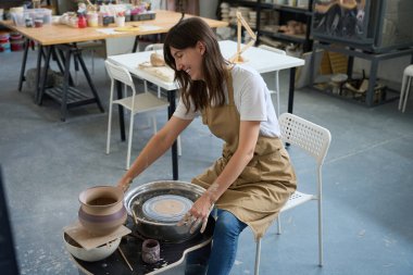 Koruyucu önlüklü ve kot pantolonlu gülümseyen bir kadın çömlekçinin yanındaki bir iş yerinde oturuyor.