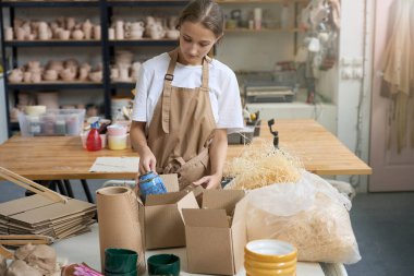 Genç bir kadın masada duruyor ve mavi bir seramik vazoyu paketlenmiş bir karton kutuya koyuyor.