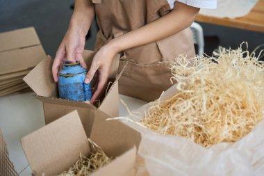 Kadın elinde mavi bir vazo tutuyor, onu bir kutuya koyup paketliyor.