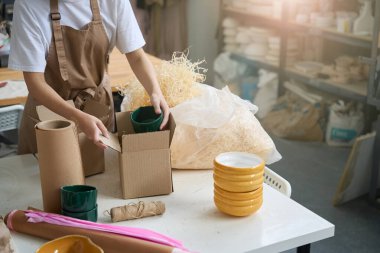 Kadın elinde yeşil bir seramik bardak tutuyor ve onu paket malzemeleriyle dolu bir kutuya koyuyor.