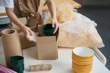 Kadın yeşil seramik bardağı karton bir kutuya koyar. Paketleme malzemesi ve sicim yakınlardadır.