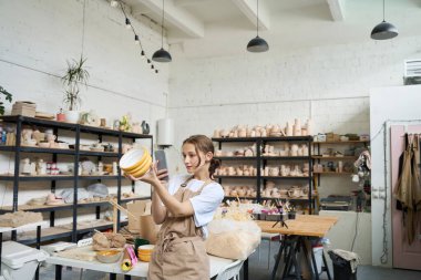 Atölyedeki öğrenci elinde dekoratif sarı vazo tutuyor ve modern bir telefonun kamerasına çekiyor.