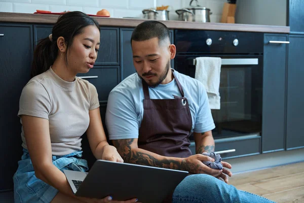 Woman shows a man something in a laptop, they are sitting on the floor in a cozy kitchen