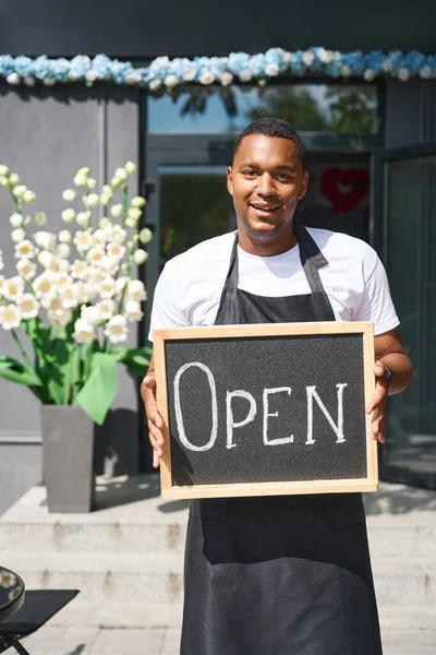 Against the backdrop of a flower shop stands a man holding a sign that says open