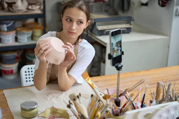 Beautiful craftswoman in an art studio sculpts plate of clay, in front of a phone camera and takes a selfie