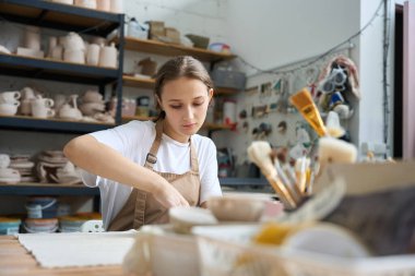 Öğrenci, yaratıcı bir atölyede arka plandaki boş çömleklerle büyük raflarda çalışıyor.