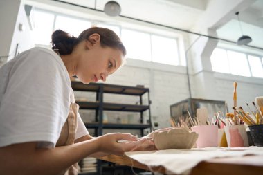 Zanaatkar kadın, masadaki kil tabağın üzerinde özel bir deste ile çalışır. Masanın üzerinde fırça ve aletlerle dolu bardaklar vardır.