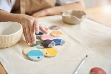 Masadaki kadının önünde kil ürünleri için yuvarlak boya örnekleri var. Elleri kille lekelenmiş.