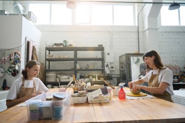 Yaratıcı stüdyoda iki kadın birbirine bakan büyük ahşap çalışma masasında oturuyor. Önlerinde boya ve fırça olan organizatörler.