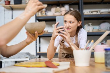 Apottery Stüdyosu 'nda iş önlüğü giyen bir öğrenci elinde bir saksıyla bir meslektaşını telefon kamerasıyla götürür.