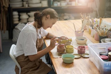 Kadın bir sanat stüdyosundaki masada oturuyor. Çamur, boya ve aletlerin arasında. Tabak boyuyor.