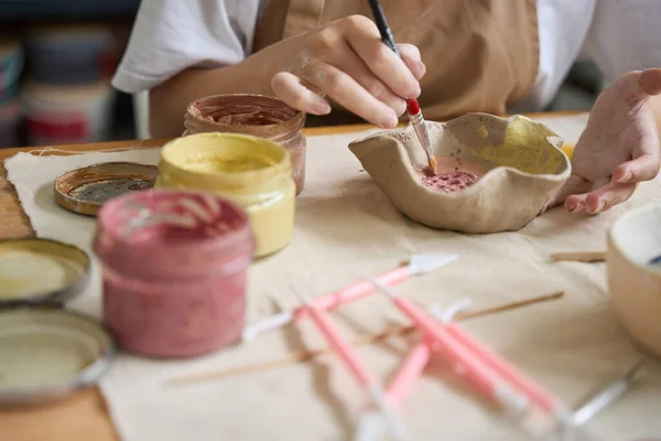 Woman paints a handmade plate with a brush, paints and tools are on the table in front of her