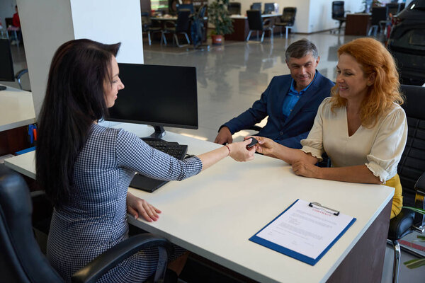 Woman manager in car dealership passes key to asatisfied client for atest drive, a clients companion sits next to her