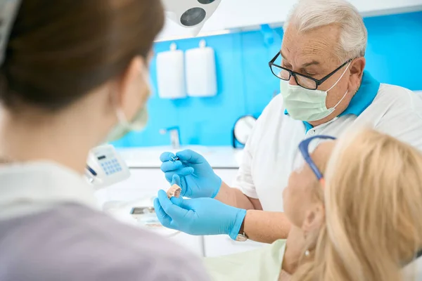 Side view portrait of elderly Caucasian dentist in blue gloves with sterile instrument in hand while pointing on the artificial teeth in dental clinic