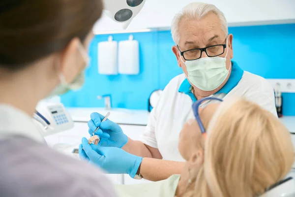 歯科医院で患者と議論しながらメガネのハンサムな男医師のクローズアップ肖像は歯モデルと金属ツールを保持しています — ストック写真