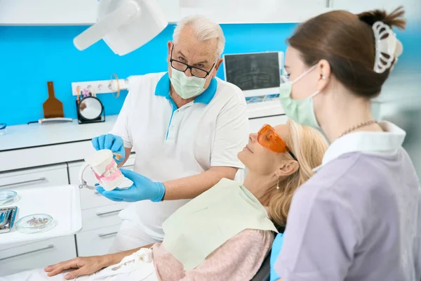 Waist up front view portrait of seriously dentist wit artificial jaw is demonstrating right way to clean teeth and gums in dental clinic