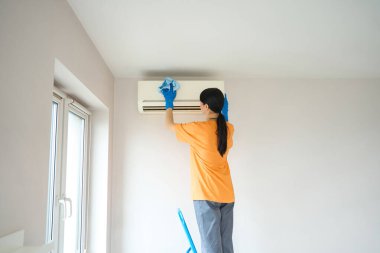 Üniformalı genç bir kadın merdivende elinde bez parçasıyla klimayı temizliyor.