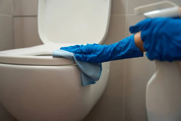 Worker holding spray for cleaning surfaces and rag, cleaning the toilet bowl in the restroom