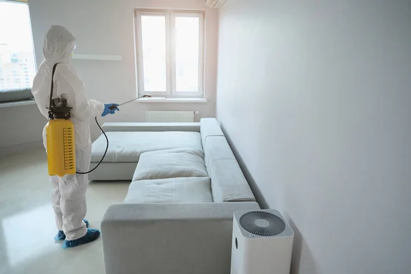 Sanitizer from cleaning company in white uniform with yellow sprayer standing in the room and disinfecting