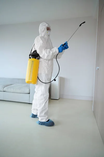 Sanitizer from cleaning company in white uniform holding yellow sprayer standing in the room and disinfecting wardrobe