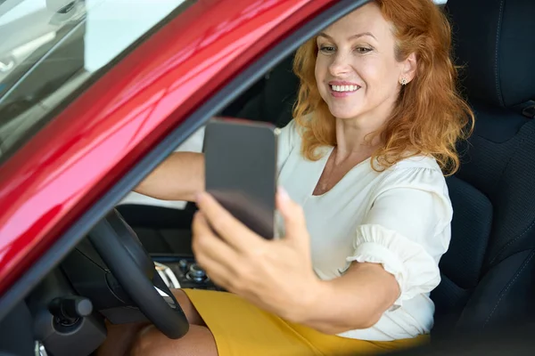 Happy woman taking photo on phone in new red auto