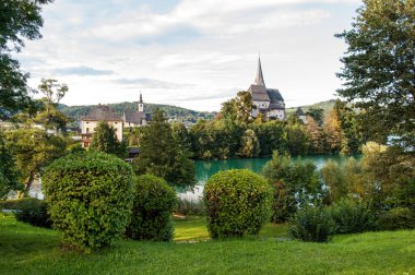 Sunlit church in the village of Maria Worth on the shores of Lake Worthersee in the Austrian mountains. Park with green grass and landscaped shrubs, a lake with crystal clear water.