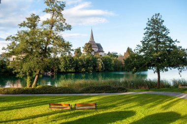 Sunlit church in the village of Maria Worth on the shores of Lake Worthersee in the Austrian mountains. Irradiated grass in the park and two benches. Beautiful lake with clear blue water.
