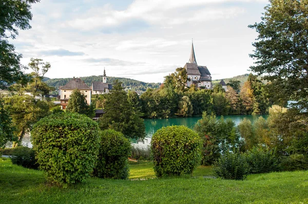 Sunlit church in the village of Maria Worth on the shores of Lake Worthersee in the Austrian mountains. Park with green grass and landscaped shrubs, a lake with crystal clear water.