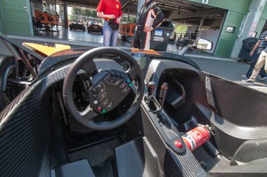 KTM yarış arabasının içi, dümende düğmeler, vites kolu ve kırmızı yangın söndürücü var. Bir arabanın önünde duran iki erkek yarışçı..