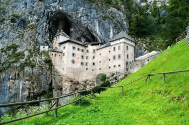 Predjama Castle in Slovenia. Old castle in the rock. Green meadow and behind it a large historic building with fortifications. clipart