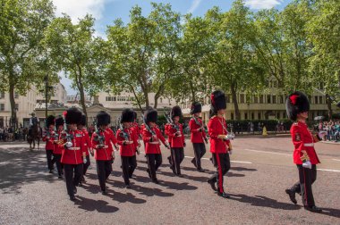 British Royal Soldiers march in a ceremony outside Buckingham Palace in central London in sunny weather. The soldiers wear red uniforms and high hats.