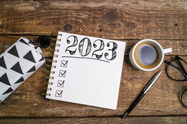 Yeni yıl hedefleri 2023 masada. Ahşap arka planda defter, kahve fincanı ve gözlüklü 2023 karar listesi. Hedefler, plan, strateji, iş, fikir, eylem konsepti