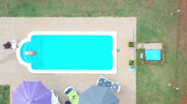 Gündüz vakti çıplak bir kadının yüzdüğü, turkuaz renkli parlak bir su ile havuz üzerine dikey drone videosu.