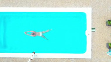 Gündüz vakti çıplak bir kadının yüzdüğü, turkuaz renkli parlak bir su ile havuz üzerine dikey drone videosu.