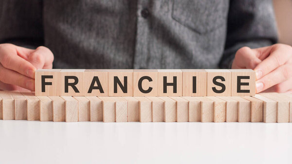 Рука кладет деревянный куб с буквой F от слова франшиза. Слово написано на деревянных кубиках, стоящих на белой поверхности стола.