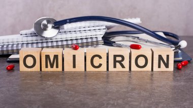 Omicron metninin yeni versiyonu gri bir masanın üzerinde duran tahta bloklardan yapılmıştır. Arka planda yazı ve tabletler için bir yığın kağıt defter var..