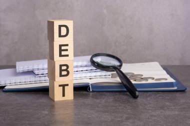 Borç kelimesi gri arka planda tahta küplere yazılır. Tahta elementlerin, büyüteçlerin, kağıt belgelerin ve bir defterin üzerindeki bozuk paraların yakın plan görüntüleri.
