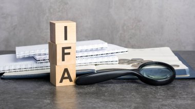 IFA kelimesi gri arka planda tahta küplere yazılır. Tahta elementlerin, büyüteçlerin, kağıt belgelerin ve bir defterin üzerindeki bozuk paraların yakın plan görüntüleri..