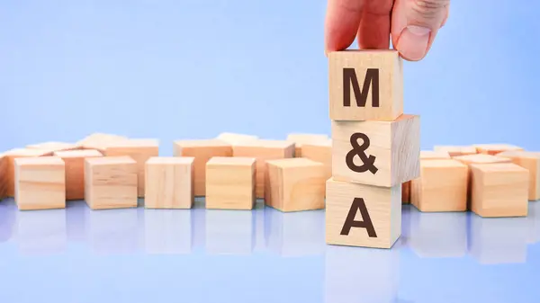 hand holding wood cube block with M and A text. the inscription on the cubes is reflected from the surface. blue background with copy space. M and A - short for Mergers and Acquisitions