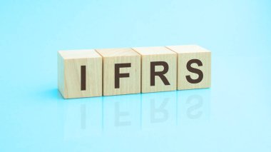 IFRS. Tahta bloklara yazılmış bir kelime. Metin siyah harflerle yazılır ve masanın ayna yüzeyine, mavi arkaplana yansıtılır. IFRS - Uluslararası Sistem Araştırmaları Federasyonu