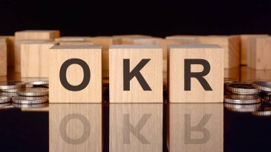 OKR - Hedefler ve Anahtar Sonuçların kısaltması. Bozuk paralar, siyah arka plan ve iş konsepti içeren tahta küplerle ilgili yazı. Küplerdeki yazıtlar siyah tablonun yüzeyinden yansıyor. ön görünüm