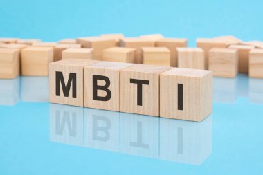MBTI. Tahta bloklara yazılmış bir kelime. Metin siyah harflerle yazılır ve masanın ayna yüzeyine, mavi arkaplana yansıtılır. MBTI - Myers-Briggs Tip Göstergesinin kısaltması